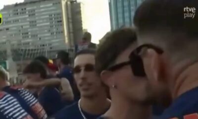 Close-up de dois homens com óculos escuros, um beijando o outro no rosto em meio à multidão
