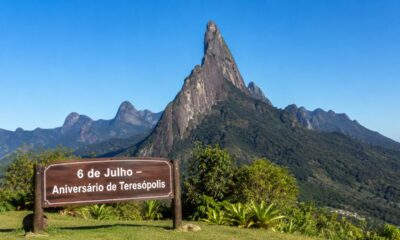 Paisagem ensolarada do Dedo de Deus, na Serra dos Órgãos, com placa em primeiro plano escrita “6 de Julho – Aniversário de Teresópolis”.