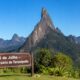 Paisagem ensolarada do Dedo de Deus, na Serra dos Órgãos, com placa em primeiro plano escrita “6 de Julho – Aniversário de Teresópolis”.