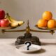 Balança de pratos equilibrada com maçãs, bananas, laranjas e pera em quantidades diferentes, com um prato à frente exibindo uma interrogação.