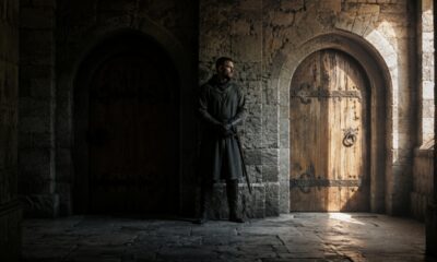Duas portas de madeira lado a lado em um castelo medieval, com um guarda entre elas e luz natural iluminando apenas uma das entradas.