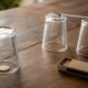 Moeda parcialmente visível sob um copo de vidro virado, com um palito apoiado entre dois copos e uma caixa de fósforos sobre uma mesa de madeira