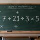 Lousa verde em uma sala de aula com a expressão “7 + 21 ÷ 3 × 5” escrita em destaque e símbolos de adição, divisão e multiplicação ao redor, sem exibir o resultado