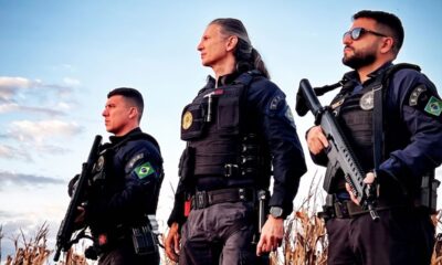 Três agentes uniformizados, com fuzis, coletes táticos e patches da bandeira do Brasil.