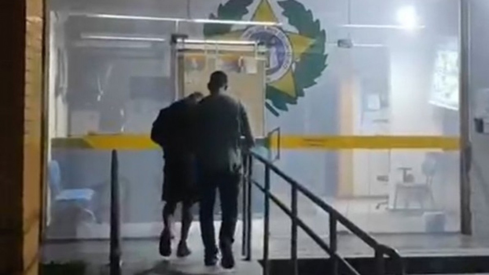 Dois homens, por trás, sobem escadas para porta de vidro com emblema policial