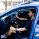Homem barbudo dirige carro azul, porta aberta. Ele segura volante e controle de marcha.