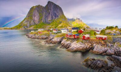 Vila costeira norueguesa com casas vermelhas sobre rochas, montanha verde e arco-íris no céu.