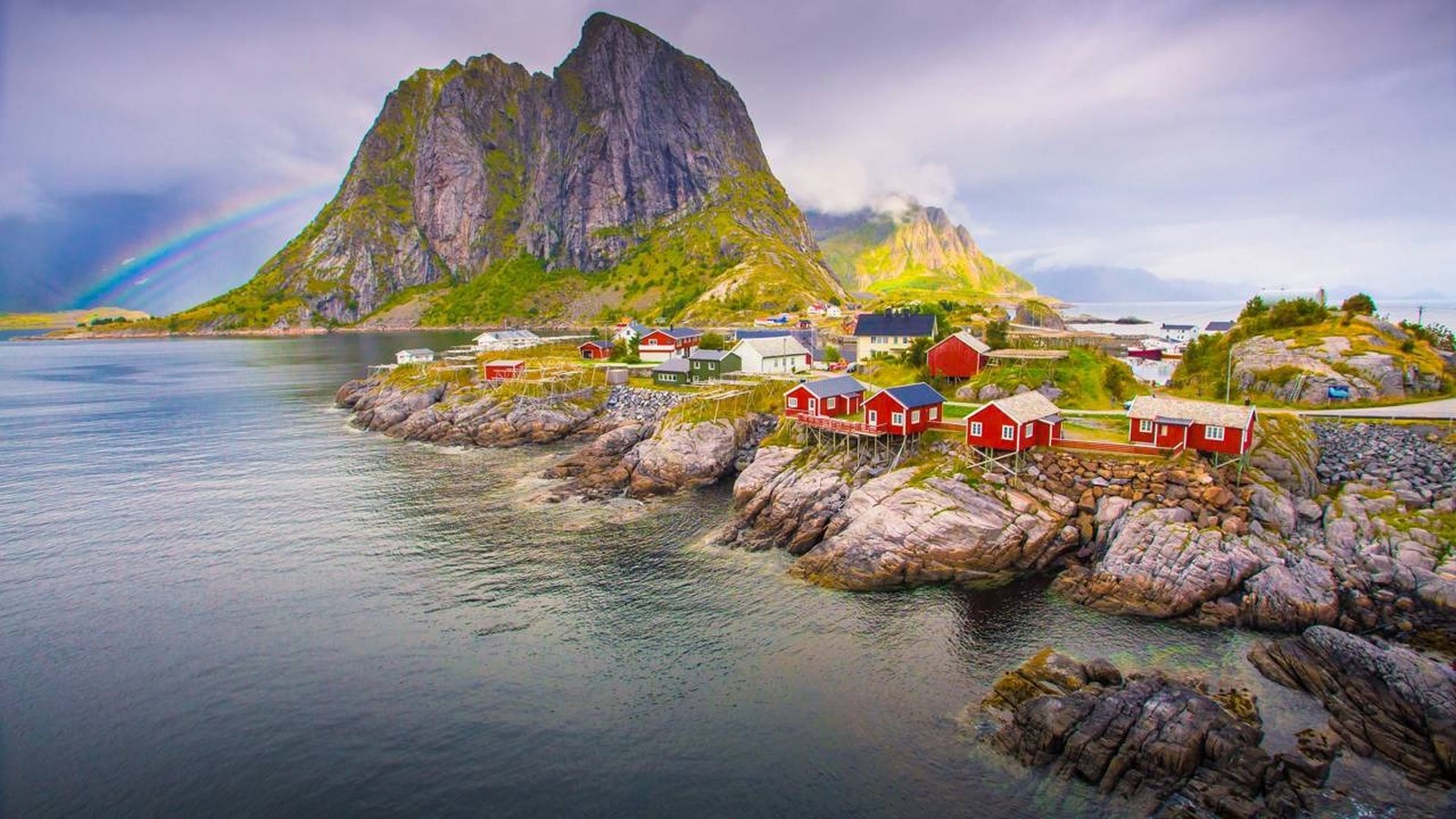 Vila costeira norueguesa com casas vermelhas sobre rochas, montanha verde e arco-íris no céu.