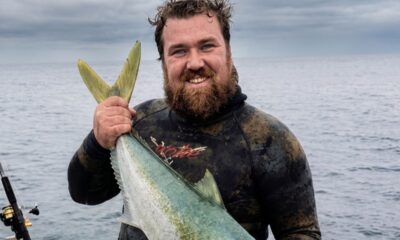 Joel Yukich, sorridente, em roupa de mergulho, segurando grande peixe sobre o mar.