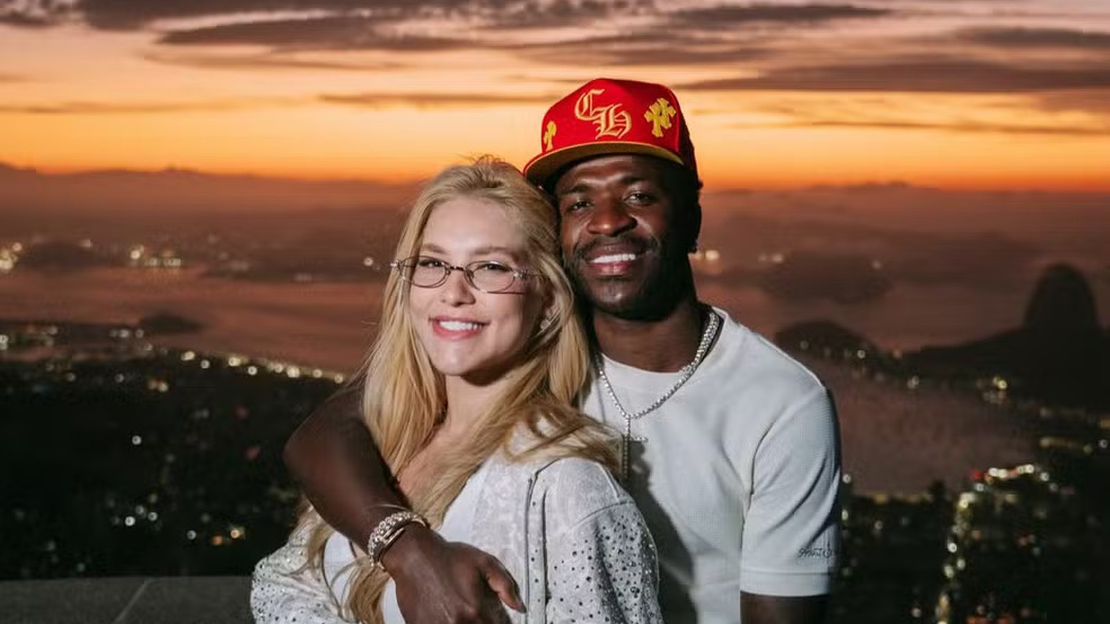 Homem e mulher sorrindo, abraçados, cidade ao pôr do sol, homem com relógios no pulso