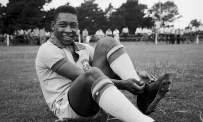 Jovem Pelé, sorrindo e sentado no gramado, ajusta chuteira com uniforme CBD