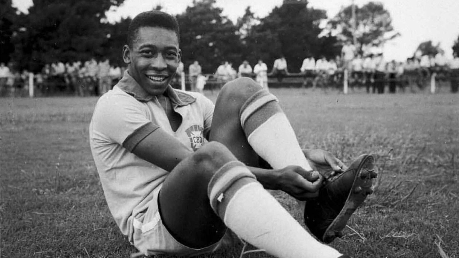 Jovem Pelé, sorrindo e sentado no gramado, ajusta chuteira com uniforme CBD