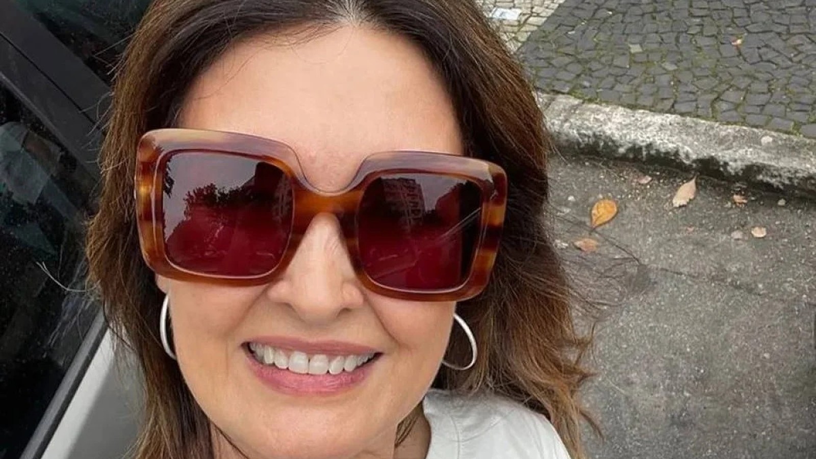 Fátima Bernardes sorrindo, com óculos de sol tartaruga quadrados e brincos de argola