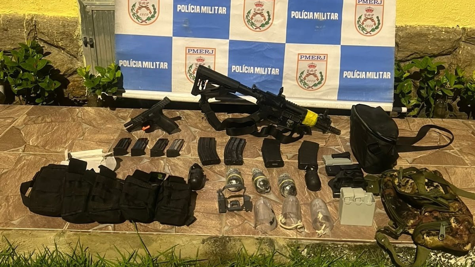 Arsenal com fuzil, pistola, granadas, carregadores e acessórios de drone sobre piso de cerâmica