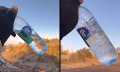 Mão enluvada segurando garrafa de água congelando sobre paisagem árida sob céu azul