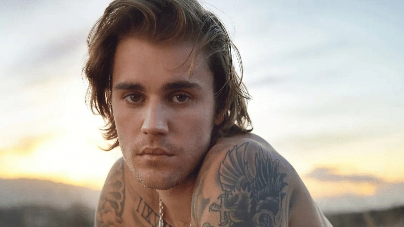 Cantor Justin Bieber sem camisa, com cabelos ondulados e tatuagens no ombro e pescoço.