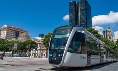 Bonde VLT cinza e preto 115 em trilhos, prédios urbanos e céu azul no Rio