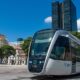 Bonde VLT cinza e preto 115 em trilhos, prédios urbanos e céu azul no Rio
