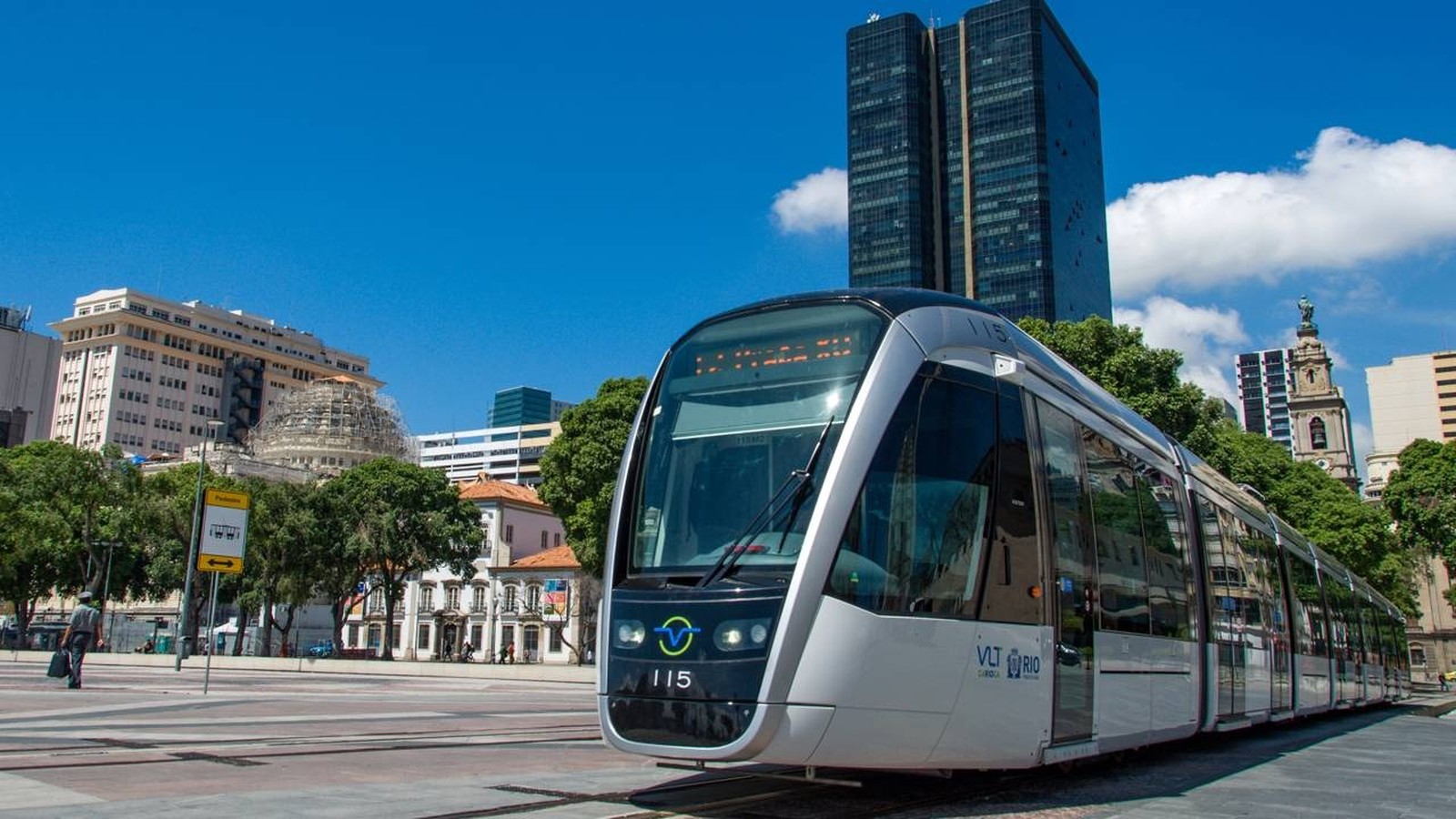 Bonde VLT cinza e preto 115 em trilhos, prédios urbanos e céu azul no Rio