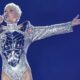 Xuxa canta com microfone, usando body prateado espelhado em palco azul