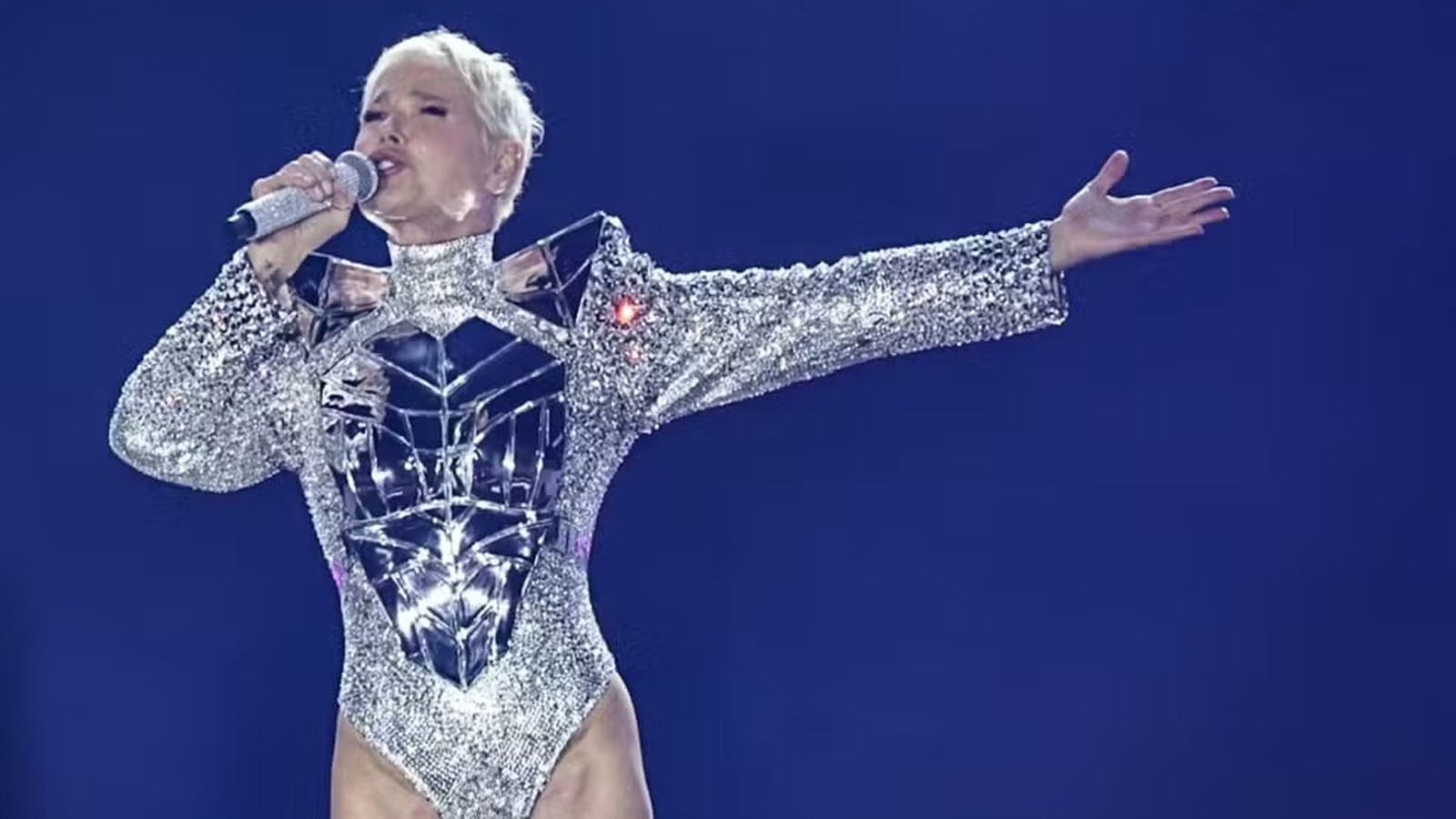 Xuxa canta com microfone, usando body prateado espelhado em palco azul