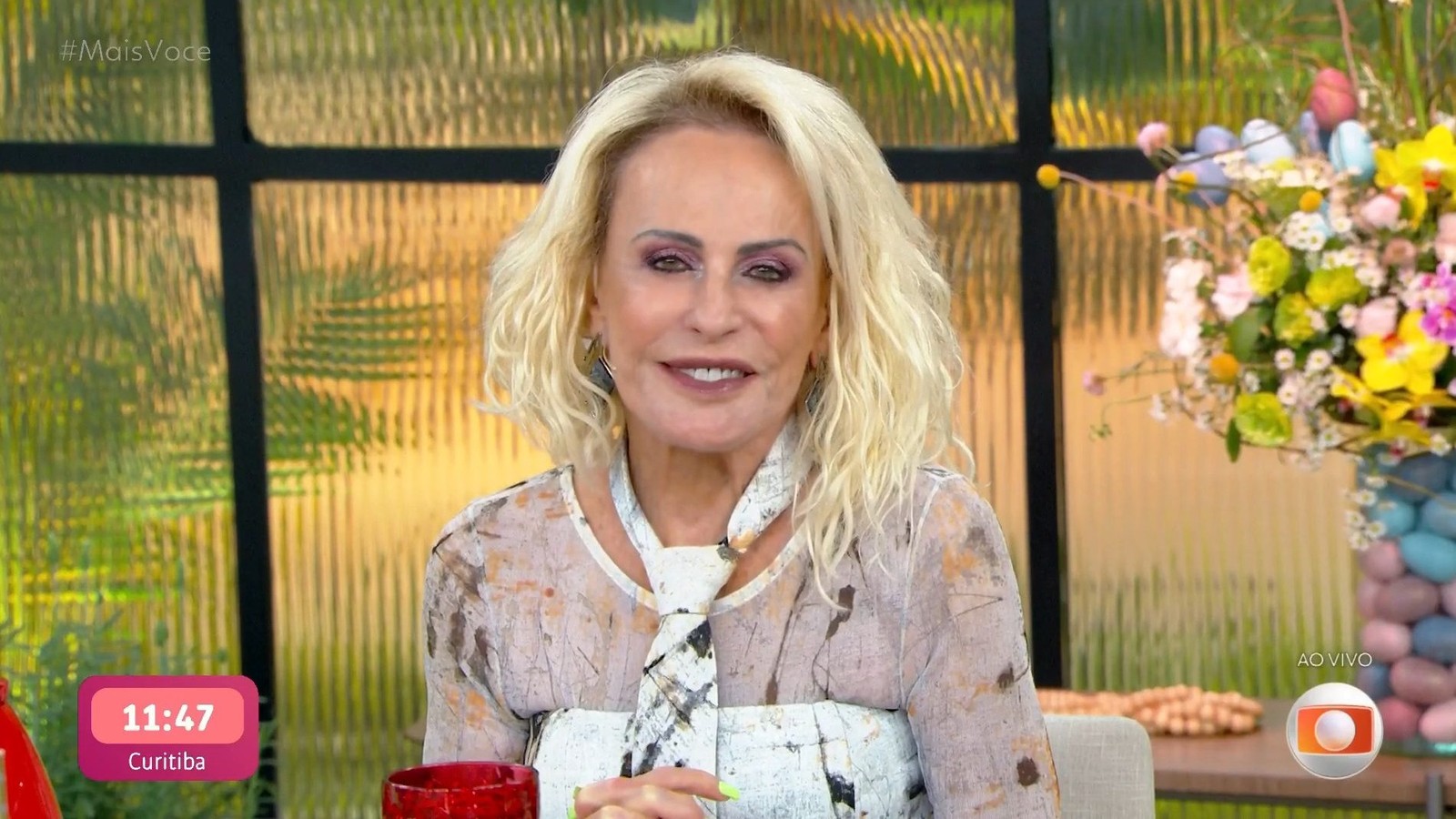 Apresentadora Ana Maria Braga sorrindo com cabelo loiro e maquiagem, exibindo "11:47 Curitiba" na tela