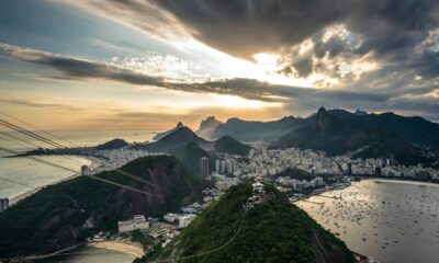 Vista panorâmica do Rio de Janeiro: montanhas, cidade, baía com barcos, e céu nublado com sol