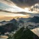 Vista panorâmica do Rio de Janeiro: montanhas, cidade, baía com barcos, e céu nublado com sol