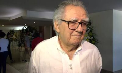 Homem idoso de óculos e cabelo grisalho em camisa branca, com arranjos florais ao fundo