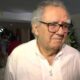 Homem idoso de óculos e cabelo grisalho em camisa branca, com arranjos florais ao fundo