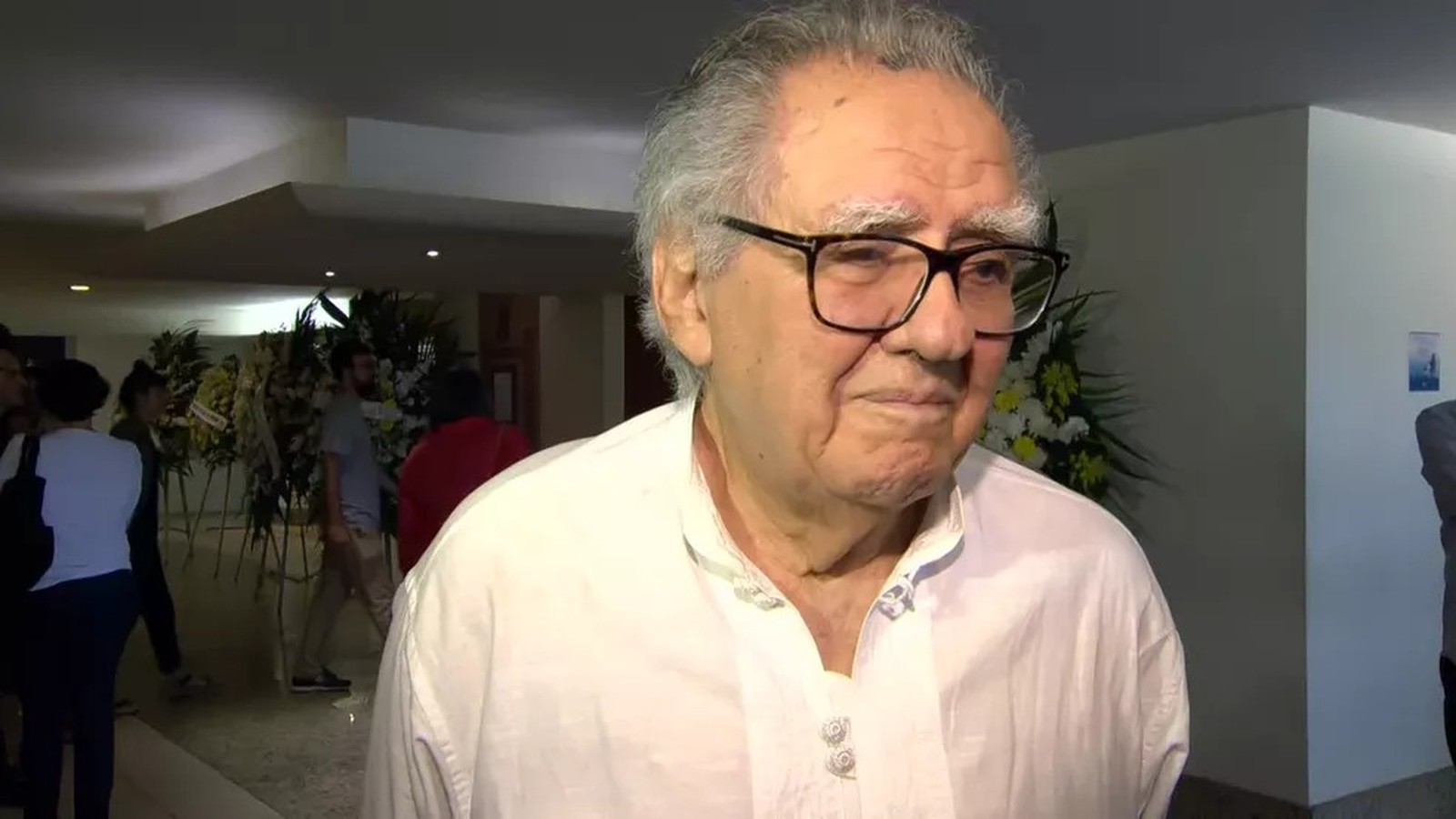 Homem idoso de óculos e cabelo grisalho em camisa branca, com arranjos florais ao fundo