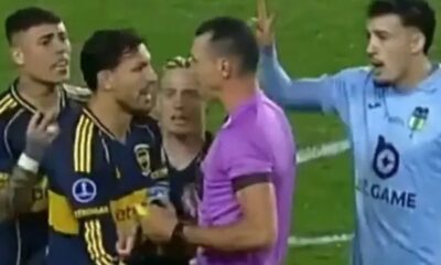 Árbitro de roxo, dois jogadores azul-amarelo e um azul-claro discutem acaloradamente em campo