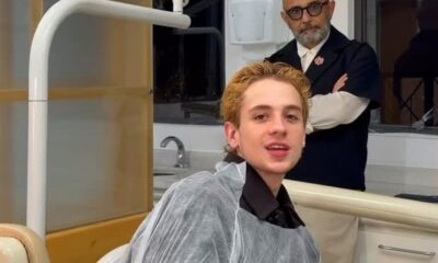 Juliano Floss em cadeira de dentista, capa protetora. Homem de óculos e barba atrás