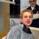 Juliano Floss em cadeira de dentista, capa protetora. Homem de óculos e barba atrás