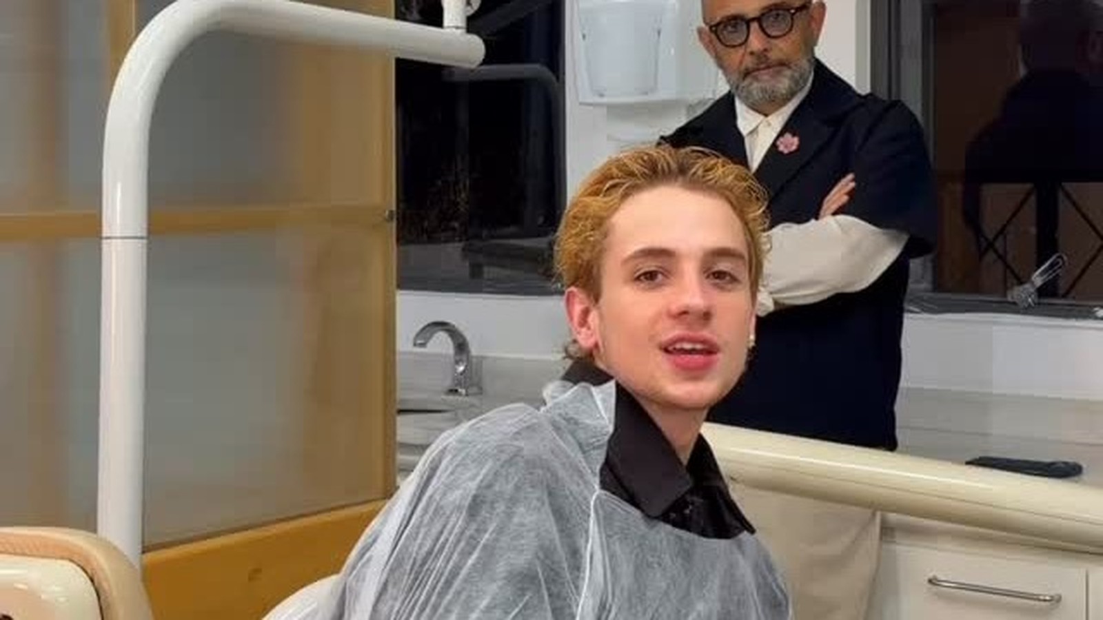 Juliano Floss em cadeira de dentista, capa protetora. Homem de óculos e barba atrás