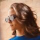 Mulher de perfil com cabelo castanho, óculos escuros e colar de pérolas, sorrindo