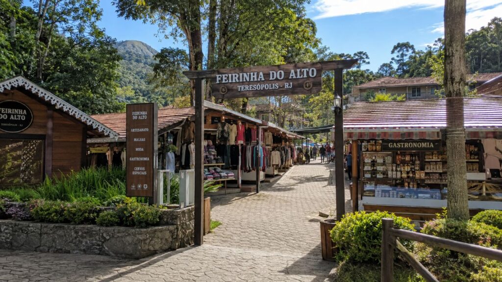 Entrada da Feirinha do Alto em Teresópolis, com barracas de roupas e gastronomia abertas, árvores ao redor, montanhas ao fundo e movimento discreto de visitantes.