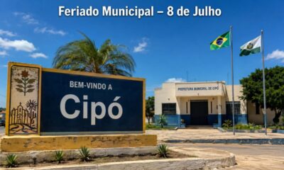 Placa de entrada de Cipó, na Bahia, com a prefeitura municipal fechada ao fundo, bandeiras hasteadas e texto no topo indicando “Feriado Municipal – 8 de Julho”, em cenário ensolarado do sertão baiano.