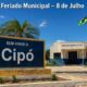 Placa de entrada de Cipó, na Bahia, com a prefeitura municipal fechada ao fundo, bandeiras hasteadas e texto no topo indicando “Feriado Municipal – 8 de Julho”, em cenário ensolarado do sertão baiano.