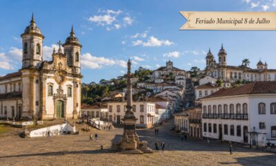 Centro histórico de Ouro Preto em dia ensolarado, com a Praça Tiradentes em destaque, igrejas barrocas, casarões coloniais nas ladeiras de pedra e faixa escrita “Feriado Municipal 8 de Julho”.
