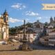 Centro histórico de Ouro Preto em dia ensolarado, com a Praça Tiradentes em destaque, igrejas barrocas, casarões coloniais nas ladeiras de pedra e faixa escrita “Feriado Municipal 8 de Julho”.