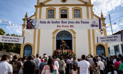 Igreja da Paróquia Nosso Senhor dos Passos, em Rio Preto (MG), com a imagem do Senhor Bom Jesus dos Passos na entrada, faixas da festa e fiéis reunidos para a celebração religiosa.