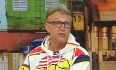 Homem de óculos e moletom estampado, com boca aberta, em um estúdio de TV colorido