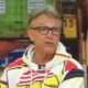 Homem de óculos e moletom estampado, com boca aberta, em um estúdio de TV colorido
