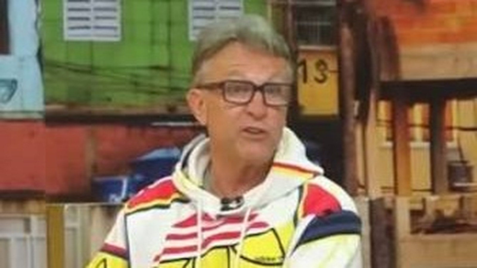 Homem de óculos e moletom estampado, com boca aberta, em um estúdio de TV colorido