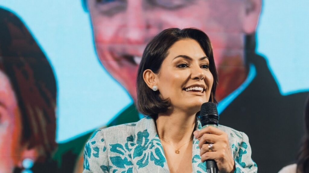 Michelle Bolsonaro sorrindo em primeiro plano, segurando microfone, com imagem de Jair Bolsonaro em fundo desfocado.