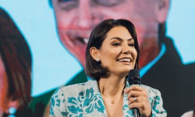 Michelle Bolsonaro sorrindo em primeiro plano, segurando microfone, com imagem de Jair Bolsonaro em fundo desfocado.