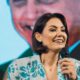 Michelle Bolsonaro sorrindo em primeiro plano, segurando microfone, com imagem de Jair Bolsonaro em fundo desfocado.