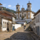 Ladeira de pedra em Ouro Preto levando até uma igreja barroca, cercada por casarões coloniais preservados, com poucos visitantes caminhando em dia ensolarado de inverno em Minas Gerais.