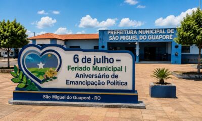 Praça central de São Miguel do Guaporé em dia ensolarado, com placa comemorativa do feriado municipal de 6 de julho e prédio da Prefeitura ao fundo.